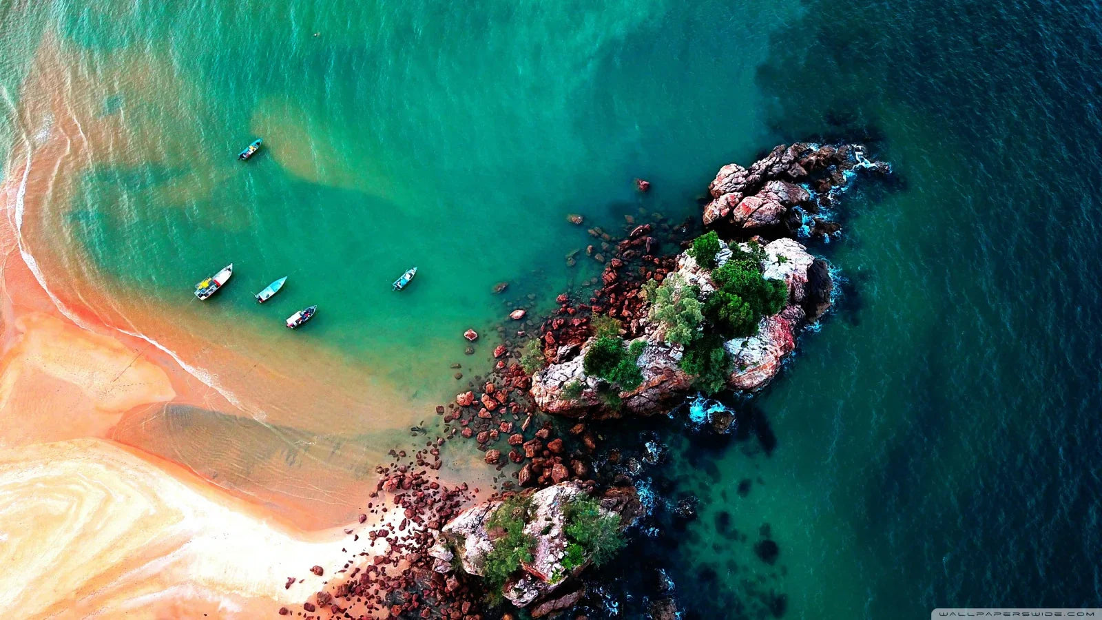 Aerial view of turquoise beach with rocky shoreline, green trees, and small boats in the water.