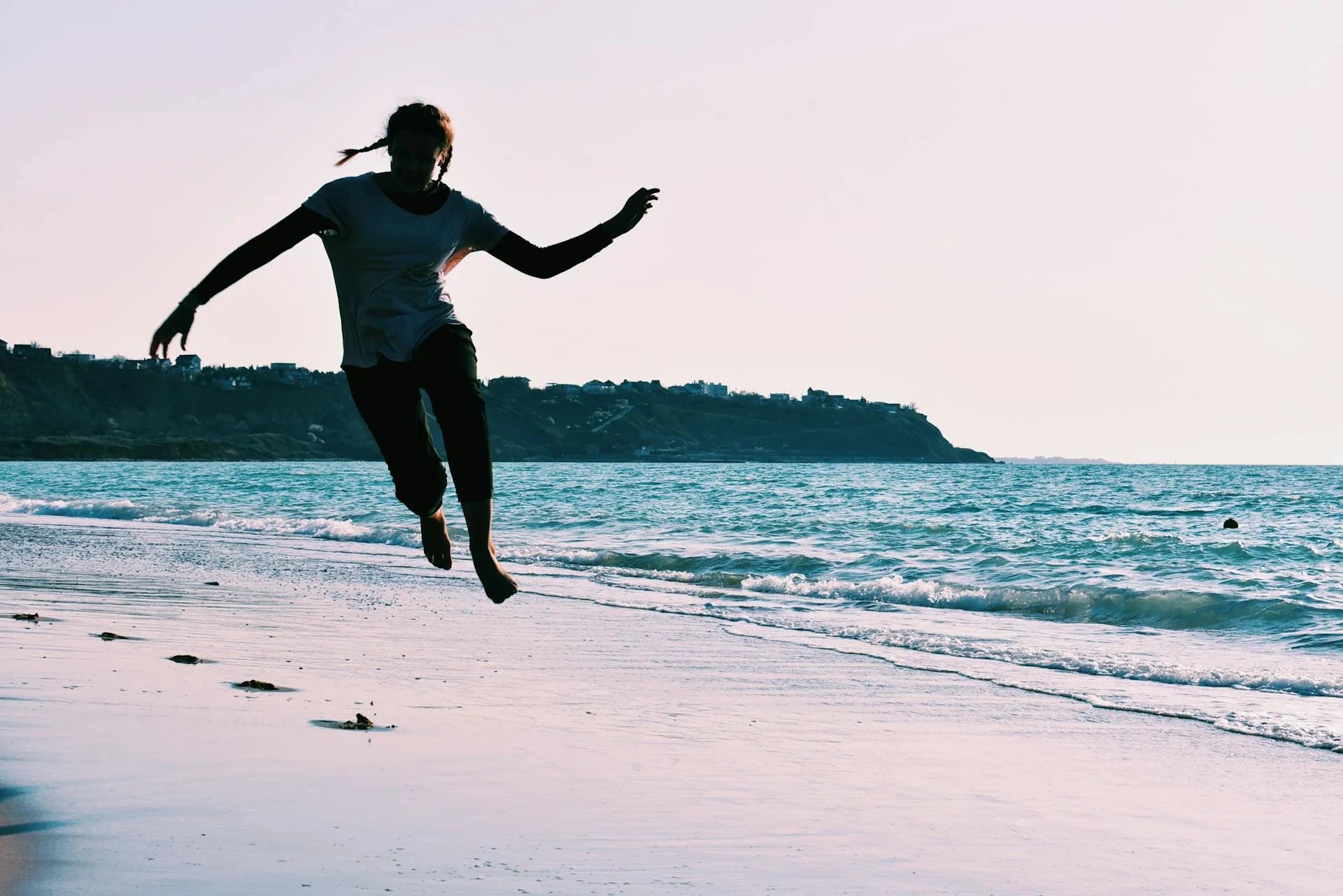 Person jumping on the beach near ocean waves, with scenic coastline in the background